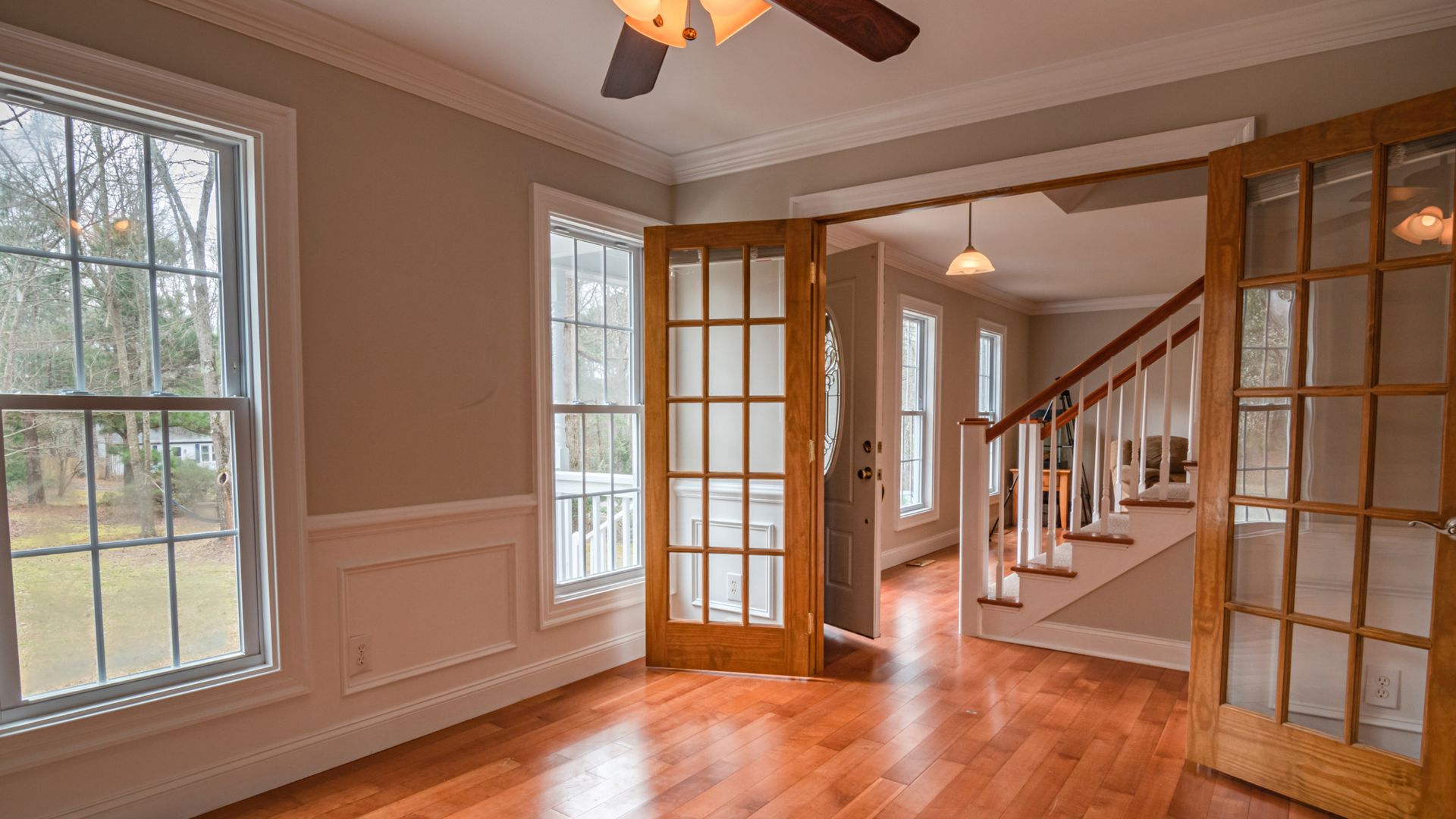 A bright room with large windows, wooden floors, and French doors—expertly crafted by a top Windows and Doors Service—leads to a hallway with stairs. Natural light fills the space, and trees are visible outside. Bramley Windows and Doors Installation and Replacement Service in Yorkshire, United Kingdom.