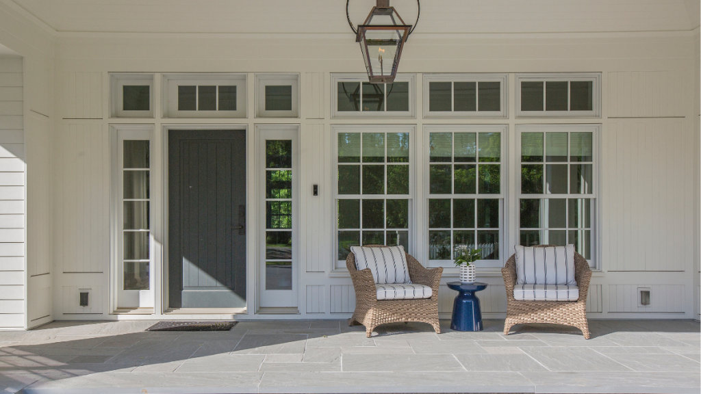A front porch with two wicker chairs with striped cushions, a small blue side table with a plant, large windows, a dark gray door, and a hanging lantern overhead. Bramley Windows and Doors Installation and Replacement Service in Yorkshire, United Kingdom.