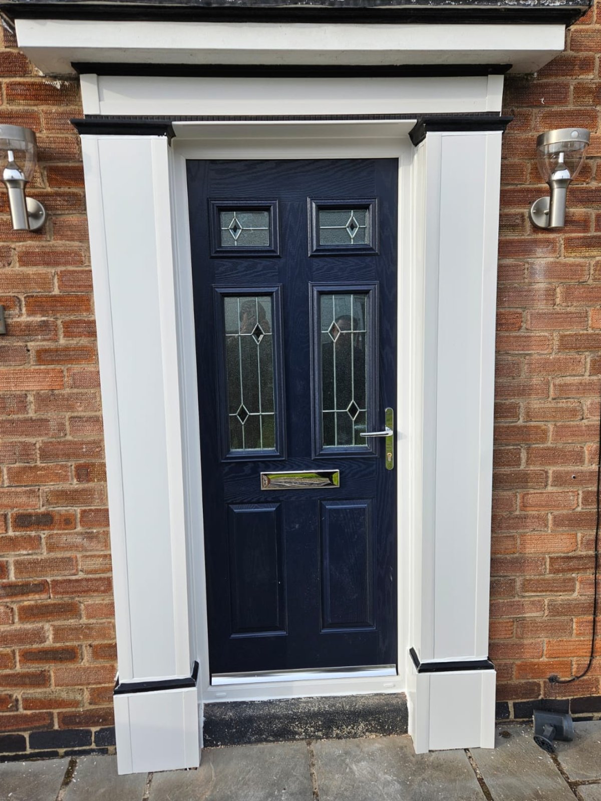 A dark blue front door with four glass panels and a gold mail slot, framed by white trim, set in a brick wall with two silver outdoor lights on either side. Bramley Windows and Doors Installation and Replacement Service in Yorkshire, United Kingdom.