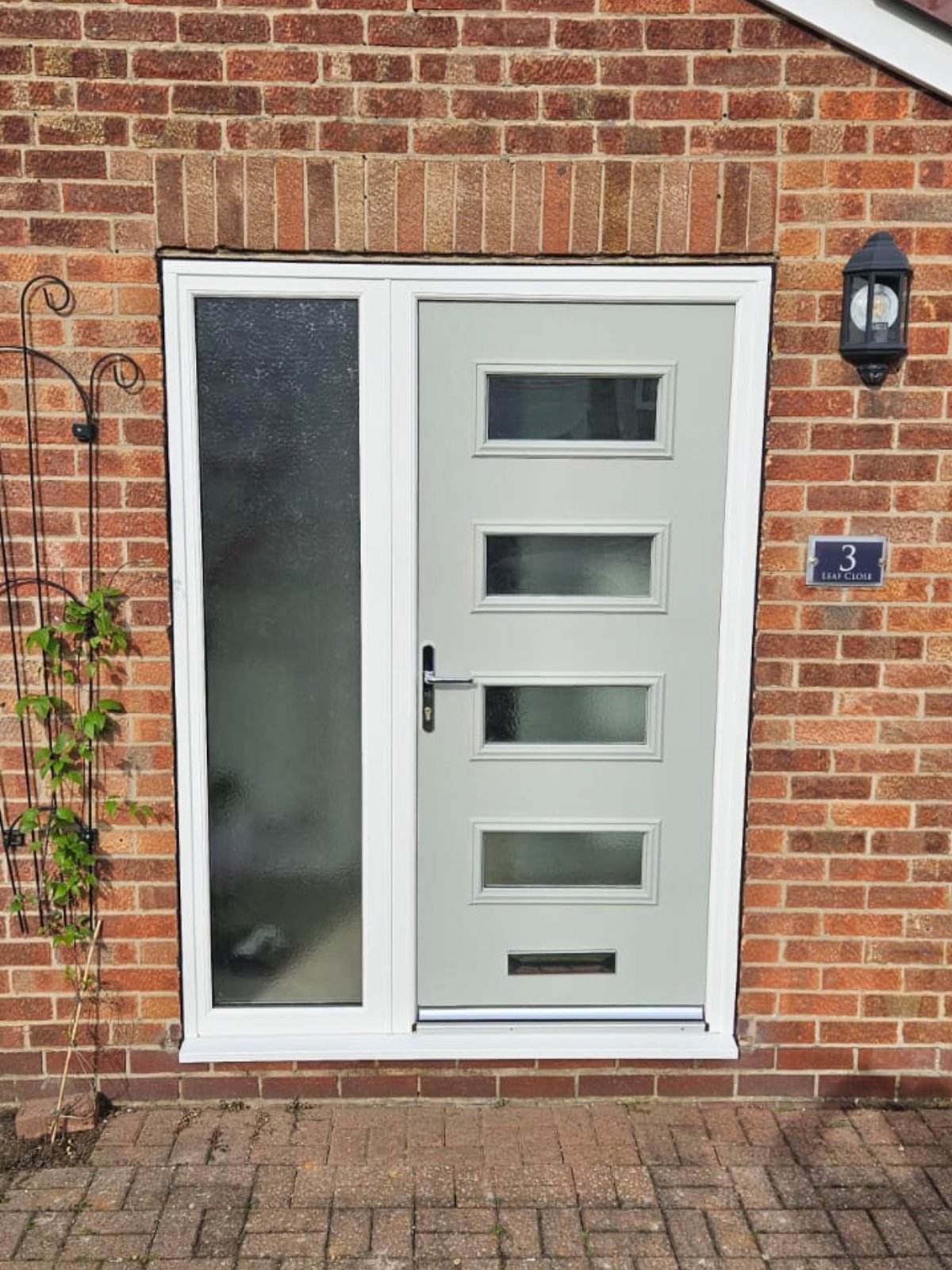 A modern light gray Completed Composite Door with four rectangular glass panels is set in a red brick wall. A frosted glass sidelight sits on the left, with a wall lantern and house number 3 plaque mounted nearby. Bramley Windows and Doors Installation and Replacement Service in Yorkshire, United Kingdom.